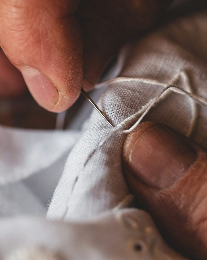 Finger führen eine Nadel mit weißem Faden durch hellen Leinenstoff