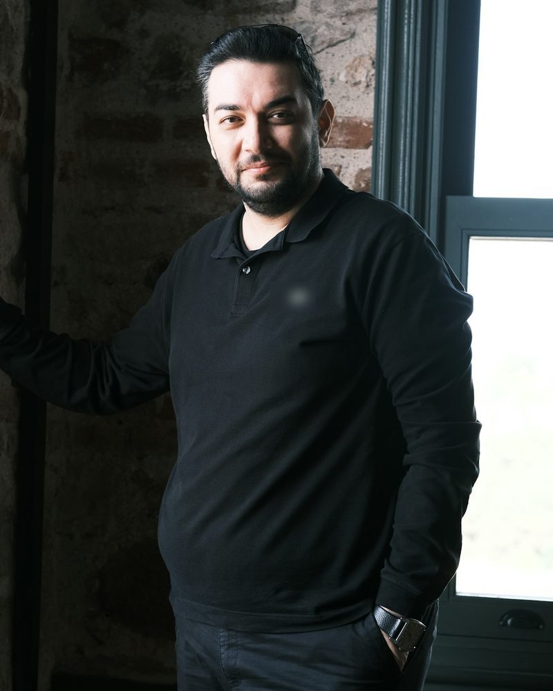Portrait von Emre mit Bart und schwarzem Poloshirt an einem Fenster vor einer Backsteinwand